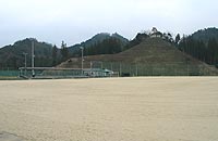 小高い山々が連なる自然豊かな風景を背景に、野球用のベンチや照明が設置された砂地のグラウンド場の写真