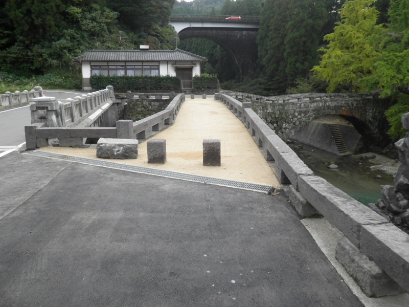 緑色に生い茂った木々を背景に道路脇に建てられた石造りの欄干とクリーム色に舗装がされた橋の先にはもう一つの石畳の橋が見えている第二二俣橋を橋の上から撮影した写真