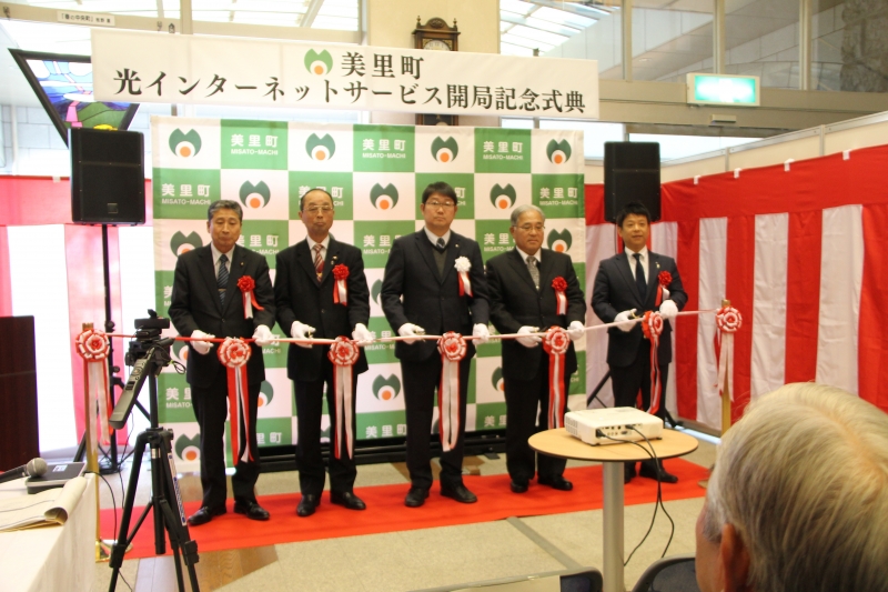 「美里町 光インターネットサービス開局記念式典」の横断幕が掲げられ、紅白の幕で囲まれたスペースで、美里町のバックボードの前に並んだ上田町長他4名の関係者が、手前の紅白のテープにハサミを入れようとしている、テープカットの記念写真