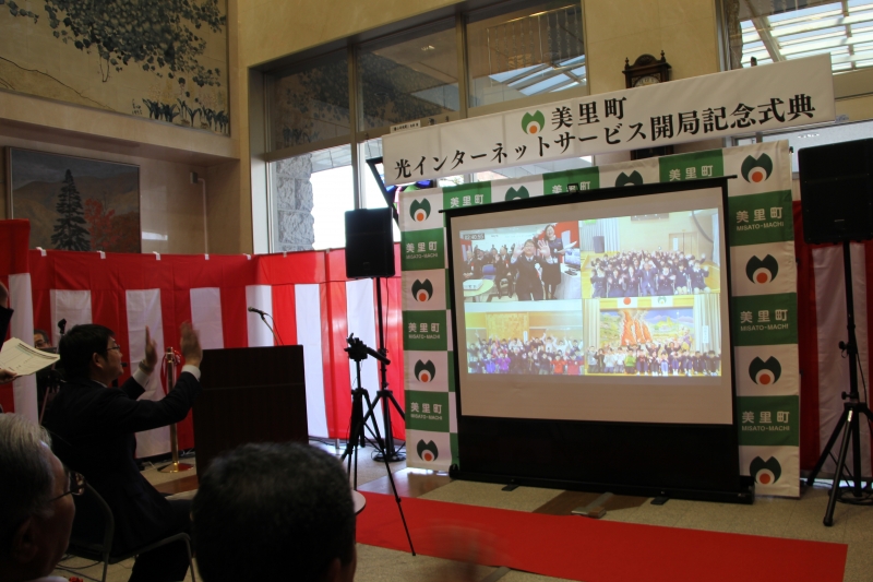 美里町のバックボード前に設置された大きなスクリーンに、手を振る上田市長の映像、3つの小学校の映像が映し出されており、会場では上田市長がスクリーンの前で手を振っている、テレビ会議の様子の写真